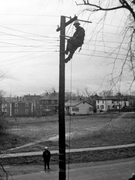 grayscale photograph: lineman atop telephone pole making repairs, empty lot in background