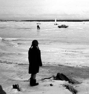 grayscale photograph: youngster viewing automobiles, pedestrians and iceboat on frozen lake