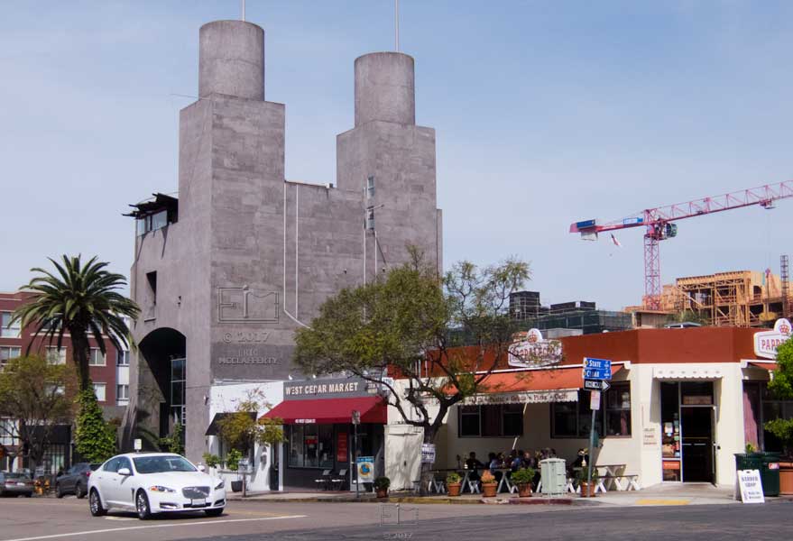 View of sidewalk restaurant / small market in front of large rectangular unpainted concrete structure topped by 2 large cylinders