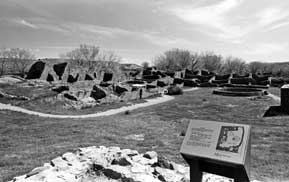 B/W photo: Sign in foreground of wide view of ruins and kivas