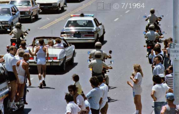Color photo: Spectators view/applaud passing torch bearer and motercade  - Embedded text: 7/1984