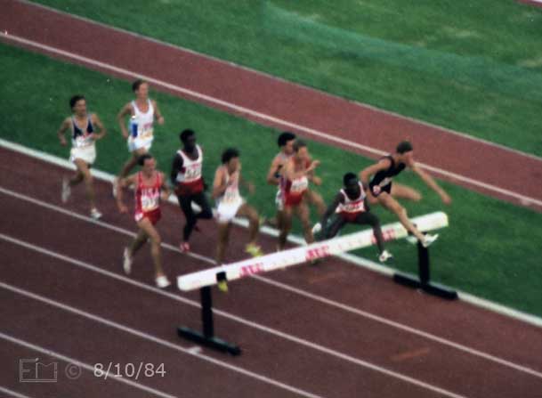 Color photo: Two steeple racers jumping hurdle with 7 others close behind  - Embedded text: 8/10/84