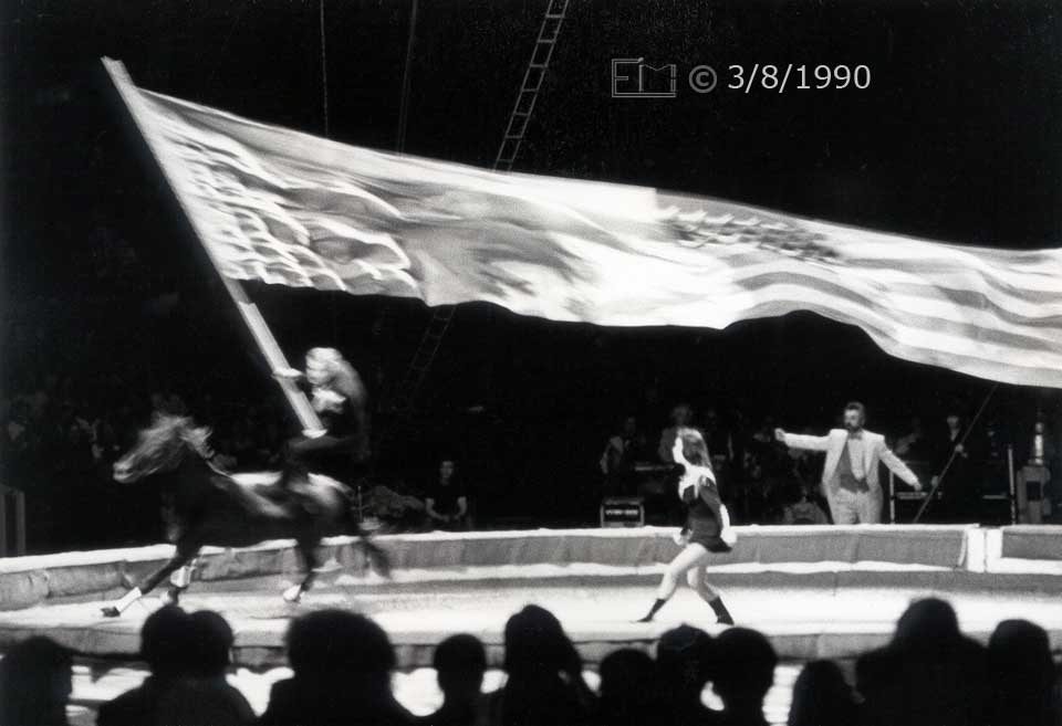 B/W photo: Performer standing on back of galloping horse holding giant Soviet/US flag - Embedded text: 3/8/1990