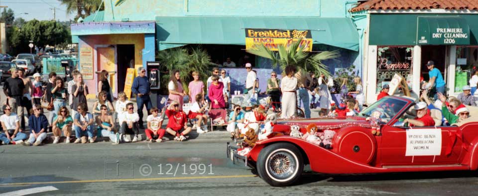 Color photo: A large, red, decorated convertible automobile passes spectators on curbside - Embedded text: 12/1991