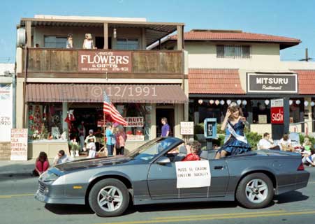 Color photo: Miss El Cajon waves to spectators from modern convertible automobile with Garnet St.  store fronts in background - Embedded text: 12/1991