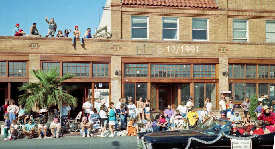 Color photo: Curbside and rooftop spectators view passing automobile - Embedded text: 12/1991