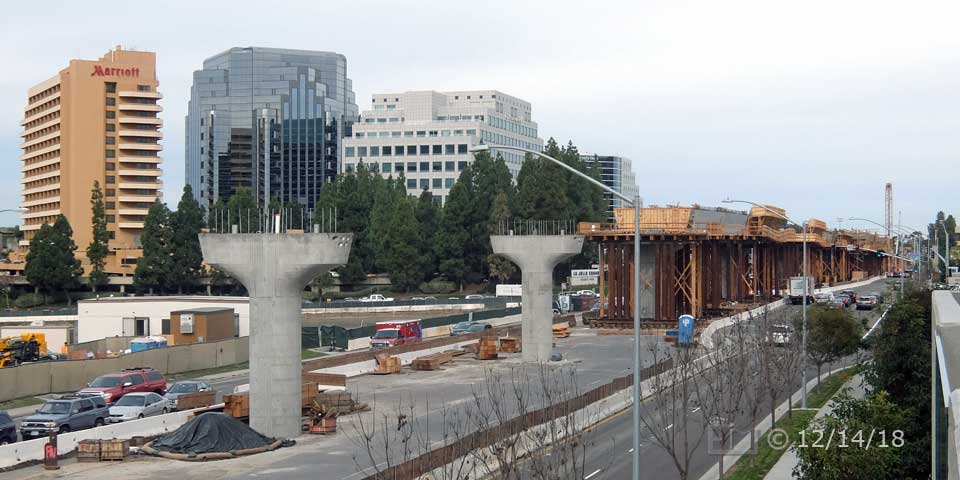 Color photo: Elevated view of support pillars and trolley line construction along Genesee Ave median strip - Embedded text: 12/14/18
