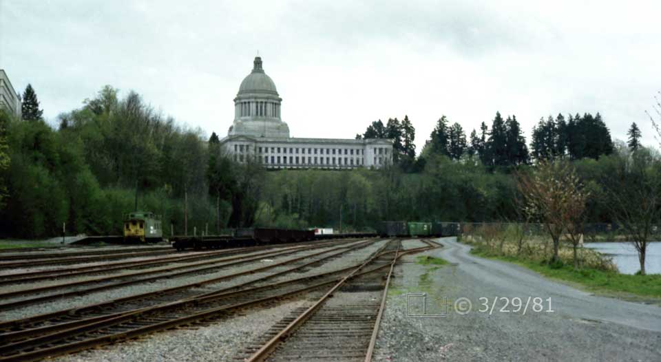Color photo: Many railway tracks in foreground with trees and back of State Capital building in background - Embedded text: 3/29/81