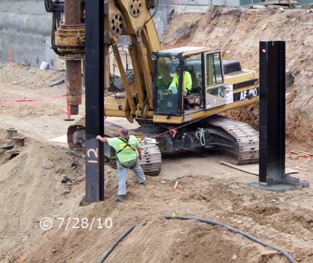 Color photo: Workman and crane operator positioning suspended I-beam above open posthole - Embedded text: 7/28/10