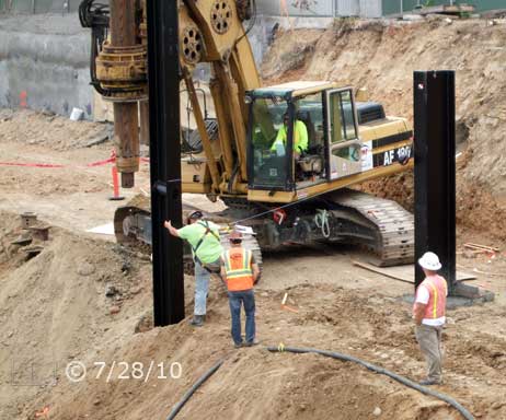 Color photo: Workman and crane operator loading suspended I-beam into open posthole - Embedded text: 7/28/10