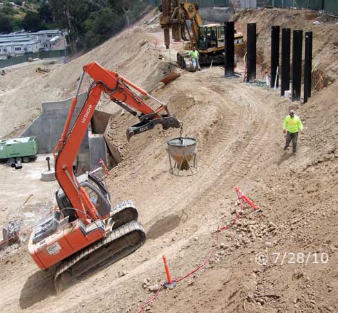 Color photo: Tracked excavator transporting loaded concrete bucket up steep incline to top of hillside  - Embedded text: 7/28/10