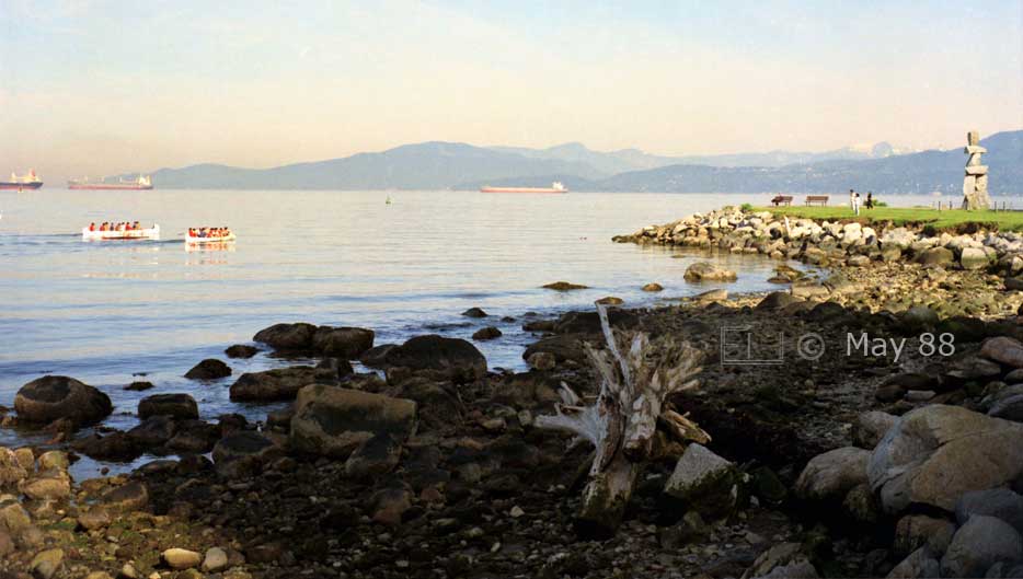 Color photo: Wide view of English Bay shore with Inukshuk Stone Sculpture on right and 2 large canoes and freighters on left - Embedded text: May 1988