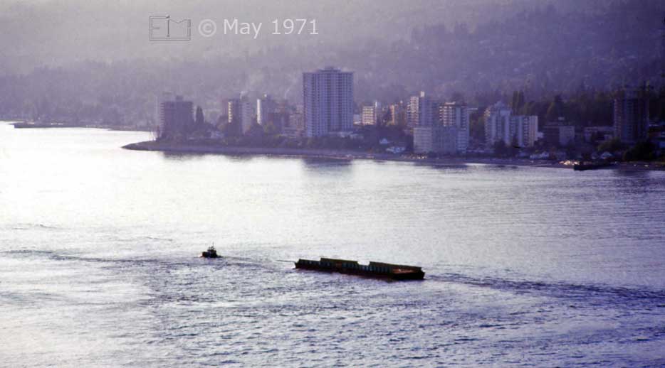 Color photo: Tug-boat and barge passing through Burrard Inlet with North Vancouver in background - Embedded text:May 1971