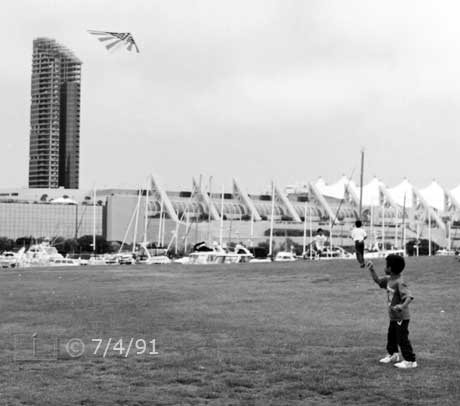 B/W photo: Boy flying kite with marina and convention center in background - Embedded text: 7/4/91