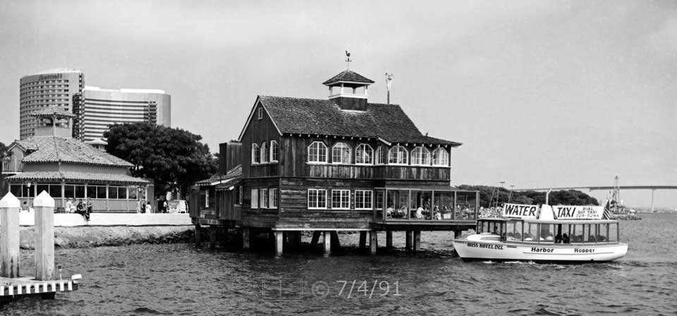 B/W photo: The 'WATER TAXI' pedestrian ferry navigates past a Seaport Village restaurant to dock - Embedded text: 7/4/91