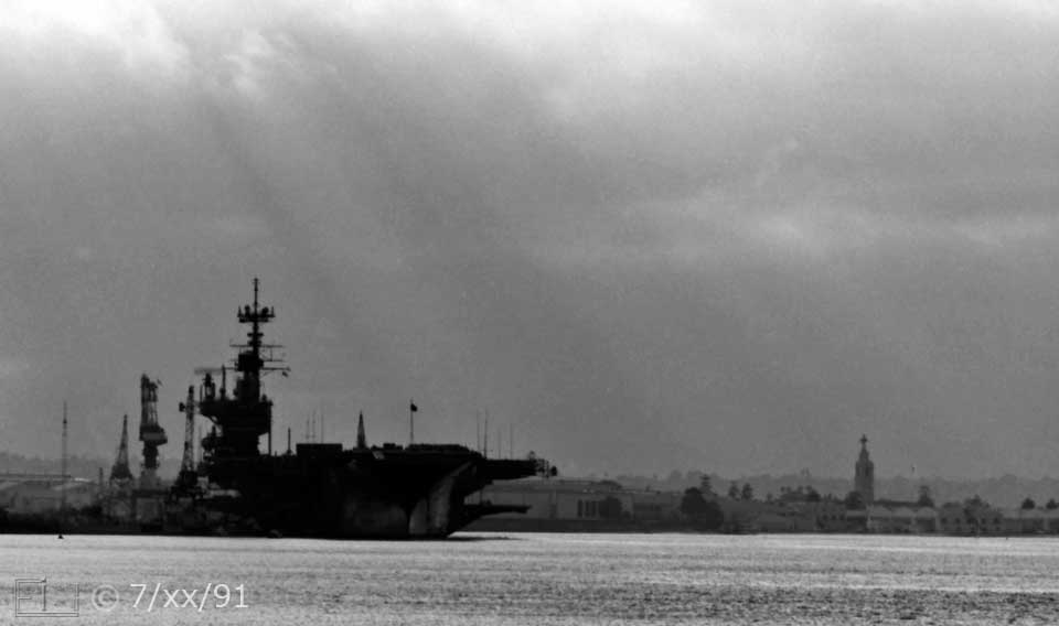 B/W photo: A docked aircraft carrier from/on Coronado side of bay - Embedded text: 7/xx/91